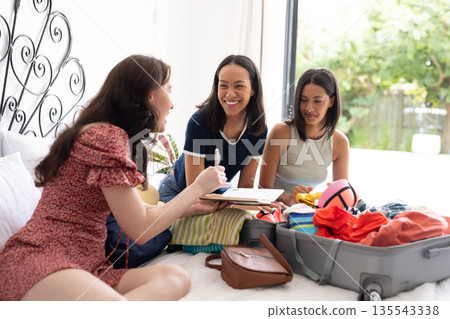 Packing suitcase on sunny day, three young women share joyful moment Packing suitcase on sunny day, three young women share joyful moment 135543338