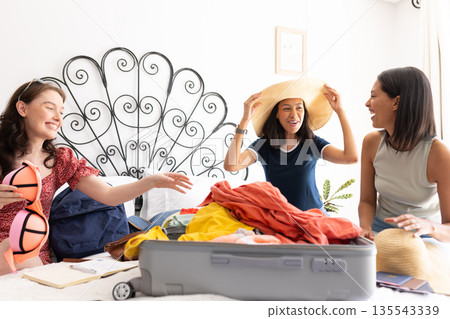 Packing suitcase with colorful clothes, three young women share joyful moment Packing suitcase with colorful clothes, three young women share joyful moment 135543339