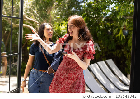 Two young women, one with long brown hair in red dress, happily entering sunlit patio 135543342