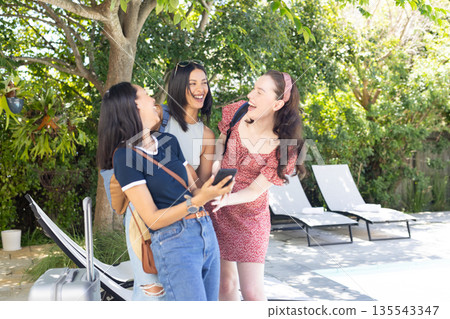 Laughing together by pool, three young women enjoy sunny day outdoors 135543347