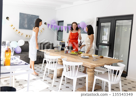 Chatting and laughing, three young women gather around wooden table at home Chatting and laughing, three young women gather around wooden table at home 135543349