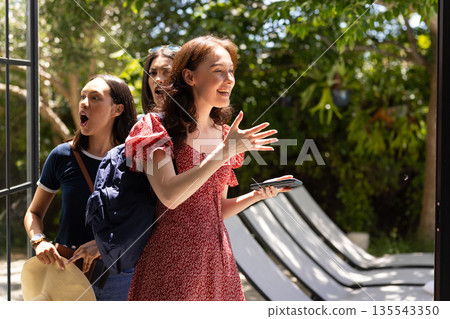 Three young women entering sunny garden, one holding smartphone and smiling Three young women entering sunny garden, one holding smartphone and smiling 135543350