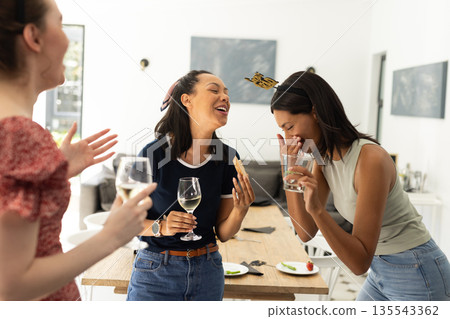 Laughing and enjoying drinks, three young women gather in bright living room Laughing and enjoying drinks, three young women gather in bright living room 135543362