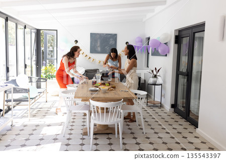 Setting up festive table, three women in colorful dresses prepare for celebration Setting up festive table, three women in colorful dresses prepare for celebration 135543397