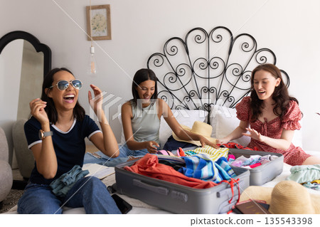 Packing suitcase and laughing, three young women enjoy sunny afternoon at home Packing suitcase and laughing, three young women enjoy sunny afternoon at home 135543398