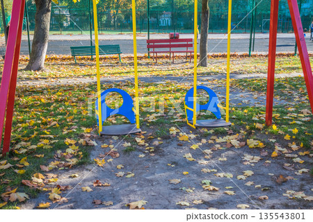 Children's swing on the playground. Yellow leaves in the playground. Gold autumn. Yellow leaves are scattered on the playground on a sunny day 135543801