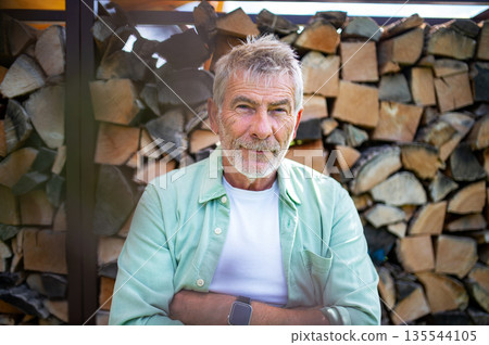 Portrait of senior man standing outdoors near stacked firewood. 135544105