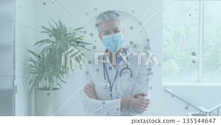 Standing doctor wearing surgical mask and button-up shirt, crossing arms in clinic with stethoscope Standing doctor wearing surgical mask and button-up shirt, crossing arms in clinic with stethoscope 135544647
