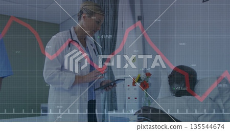 Reviewing doctor in lab coat with stethoscope scrolling tablet in ward with bed, curtain, flowers 135544674