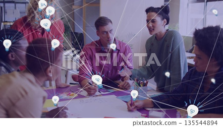 Discussing team of six leaning over sketches at meeting table in office, with lightbulb overlays 135544894