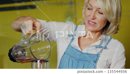 Home cook wearing denim apron pouring red smoothie from blender into container at kitchen counter Home cook wearing denim apron pouring red smoothie from blender into container at kitchen counter 135544936