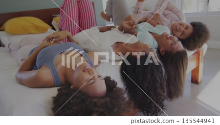 Laughing four friends wearing pajamas lying on bed in bedroom, with white linens and yellow pillows Laughing four friends wearing pajamas lying on bed in bedroom, with white linens and yellow pillows 135544941
