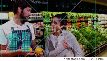 Talking employee wearing striped apron and shopper selecting orange in produce area with signage 135544982