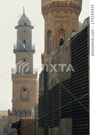 Historic Qalawun Complex in Cairo, featuring intricate architecture, tall minarets, and domes under a blue sky with clouds. A significant cultural landmark on Al-Muizz Street. 135545935
