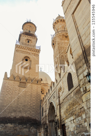 Historic Qalawun Complex in Cairo, featuring intricate architecture, tall minarets, and domes under a blue sky with clouds. A significant cultural landmark on Al-Muizz Street. Historic Qalawun Complex in Cairo, featuring intricate architecture, tall minarets, and domes under a blue sky with clouds. A significant cultural landmark on Al-Muizz Street. 135545946