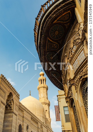 Historic Qalawun Complex in Cairo, featuring intricate architecture, tall minarets, and domes under a blue sky with clouds. A significant cultural landmark on Al-Muizz Street. Historic Qalawun Complex in Cairo, featuring intricate architecture, tall minarets, and domes under a blue sky with clouds. A significant cultural landmark on Al-Muizz Street. 135545949