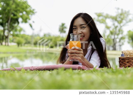 Asian Women picnic in green park smilng face hands holding glass of orange juice fresh fruit cool summer drinking. Beautiful women laydown picnic mat fruit basket hands holding healthy drinking glass Asian Women picnic in green park smilng face hands holding glass of orange juice fresh fruit cool summer drinking. Beautiful women laydown picnic mat fruit basket hands holding healthy drinking glass 135545988