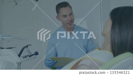 Gloved dentist wearing scrubs explaining care in treatment room, with file folder, overhead light 135546321