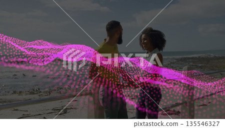 Standing couple leaning on wooden railing at beach promenade, with pink digital wave overlay 135546327