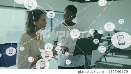 Discussing professional women sharing files on laptop in open-plan office, with overlay icons Discussing professional women sharing files on laptop in open-plan office, with overlay icons 135546379