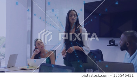 Presenting woman in blazer speaking at office meeting table, with laptops and numeric overlay Presenting woman in blazer speaking at office meeting table, with laptops and numeric overlay 135546431