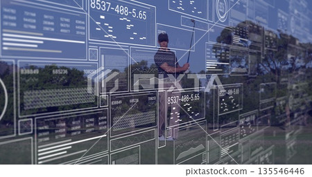 Swinging golfer gripping club and teeing golf ball on tree-lined fairway, with digital data panels 135546446