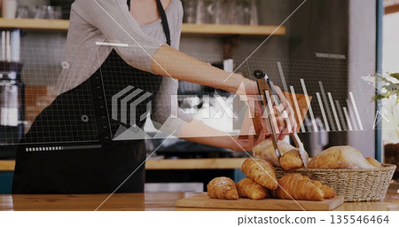 Using metal tongs barista wearing apron picking croissants behind counter at cafe with data overlay 135546464
