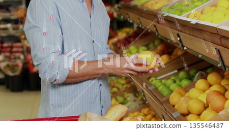shopper in striped shirt choosing orange in produce aisle with shelves, red cart holding baguettes shopper in striped shirt choosing orange in produce aisle with shelves, red cart holding baguettes 135546487