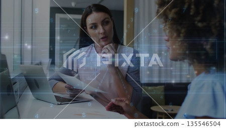 Two businesswomen discussing data on laptop and printed documents in meeting room, with pens 135546504