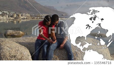 Sitting couple by lake wearing denim jacket and red top, holding smartphone under world map overlay Sitting couple by lake wearing denim jacket and red top, holding smartphone under world map overlay 135546718