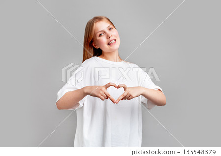 Beautiful young blond girl making modern finger heart gesture, smiling while posing in studio on clean uniform background, isolated in bright contemporary portrait, copy space 135548379