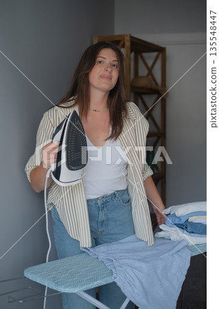 Home portrait of brunette in jeans and shirt doing morning routine preparing clothes for work day with iron Home portrait of brunette in jeans and shirt doing morning routine preparing clothes for work day with iron 135548447