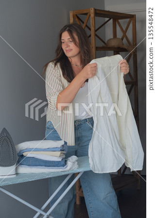 Morning home ritual with brunette carefully preparing clothes for work day holding an iron in hand 135548472