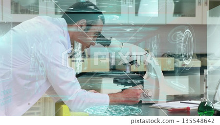 Leaning researcher wearing lab coat adjusting microscope focus in lab, with flask and hologram data 135548642