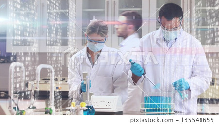Lab scientists in coats and gloves pipetting blue solution into tube rack in lab, with micropipette Lab scientists in coats and gloves pipetting blue solution into tube rack in lab, with micropipette 135548655