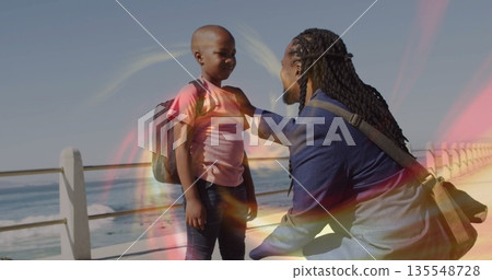 Kneeling father adjusting son's backpack strap on seaside walkway, messenger bag by railing Kneeling father adjusting son's backpack strap on seaside walkway, messenger bag by railing 135548728