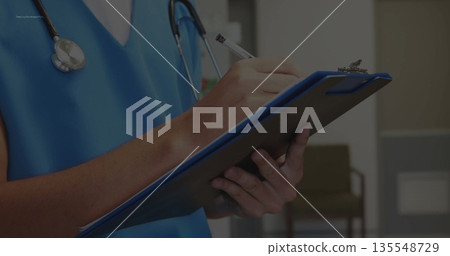 Clinician holding clipboard and writing notes in clinic hallway with scrubs and stethoscope Clinician holding clipboard and writing notes in clinic hallway with scrubs and stethoscope 135548729
