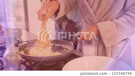 Stirring woman in bathrobe cooking scrambled eggs in kitchen as partner watches with spatula pan Stirring woman in bathrobe cooking scrambled eggs in kitchen as partner watches with spatula pan 135548749