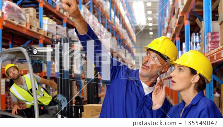 Pointing staff in blue coveralls and hard hats standing in warehouse, with forklift, chart overlay 135549042