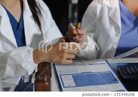Medical team having a meeting with doctors in white lab coats and surgical scrubs seated at a table discussing a patients working online using computers in the medical industry. Medical team having a meeting with doctors in white lab coats and surgical scrubs seated at a table discussing a patients working online using computers in the medical industry. 135549106