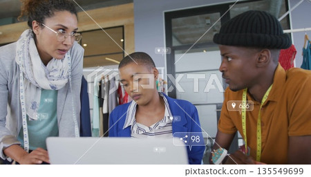Collaborating fashion designers wearing beanie at laptop in studio with measuring tapes pin cushion 135549699