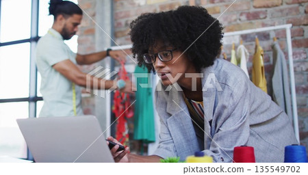 Female fashion designer leaning on desk holding stylus in loft studio, with laptop and tape measure 135549702