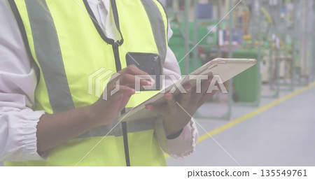 Using tablet smartphone, technician wearing safety vest inspecting factory floor, copy space 135549761