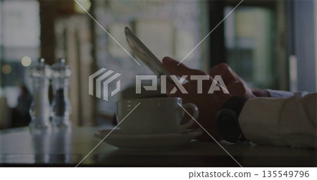 Tapping tablet, man in white shirt sitting at cafe table, with coffee cup and salt shakers Tapping tablet, man in white shirt sitting at cafe table, with coffee cup and salt shakers 135549796