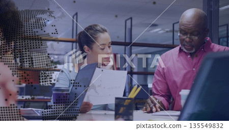 Man in pink shirt pointing with woman in blue top listening at office desk, digital overlay 135549832