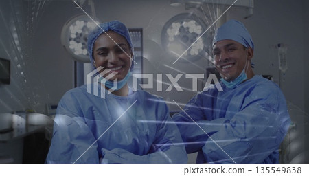 Smiling two surgeons wearing blue gowns posing in operating room, with overhead lights and monitors Smiling two surgeons wearing blue gowns posing in operating room, with overhead lights and monitors 135549838