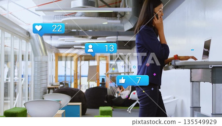 Blazer-clad woman using laptop and smartphone in office lounge, with social media icons, copy space 135549929