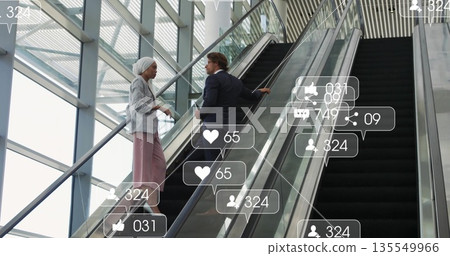 Talking business colleagues climbing escalator holding rail in glass atrium, with social icons Talking business colleagues climbing escalator holding rail in glass atrium, with social icons 135549966