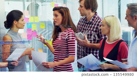 Collaborating team viewing sticky note glass wall in office with papers, tablet, phone and markers 135549980