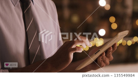 Tapping businessman in shirt and tie holding touchscreen tablet in lounge, with warm string lights 135549984
